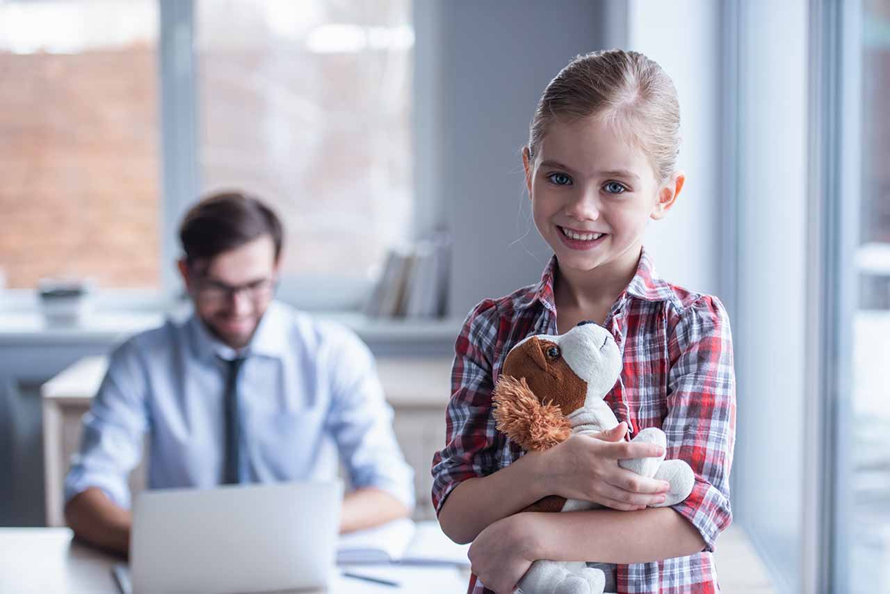 Child Support 2 Smiling Child Holding A Stuffed Toy With A Lawyer Working In The Background, Symbolising Personalised Child Support Legal Services In Melbourne And Dandenong.